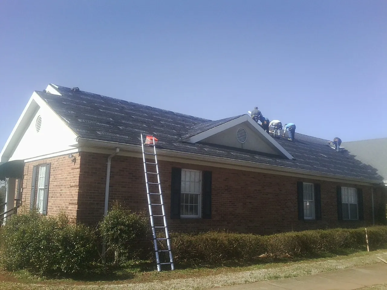 Crew tearing shingles off a brick ranch roof with a ladder set — Metal Roof Repair work by our crew in Webster